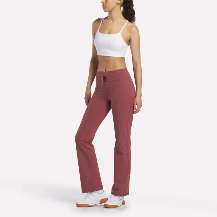 A woman stands against a white background, wearing a white sports bra, white sneakers, and mauve Reebok Women's Highstreet High-Rise Pants. She looks to the side with one hand on her hip.