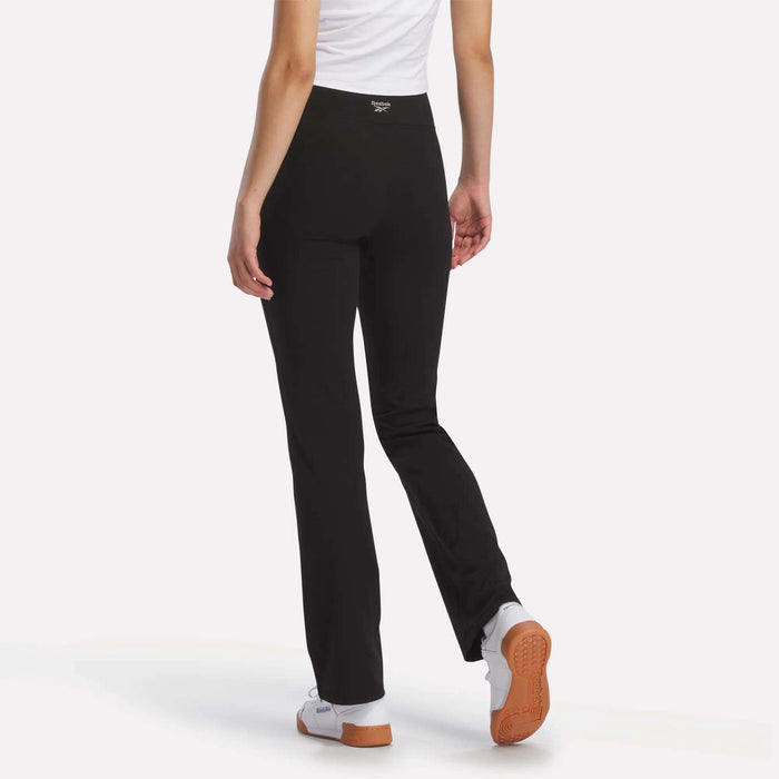 A model is shown from the back on a white background, wearing Reebok Women's Highstreet High-Rise Pants and a white shirt, paired with white and orange shoes. One arm hangs relaxed at her side.
