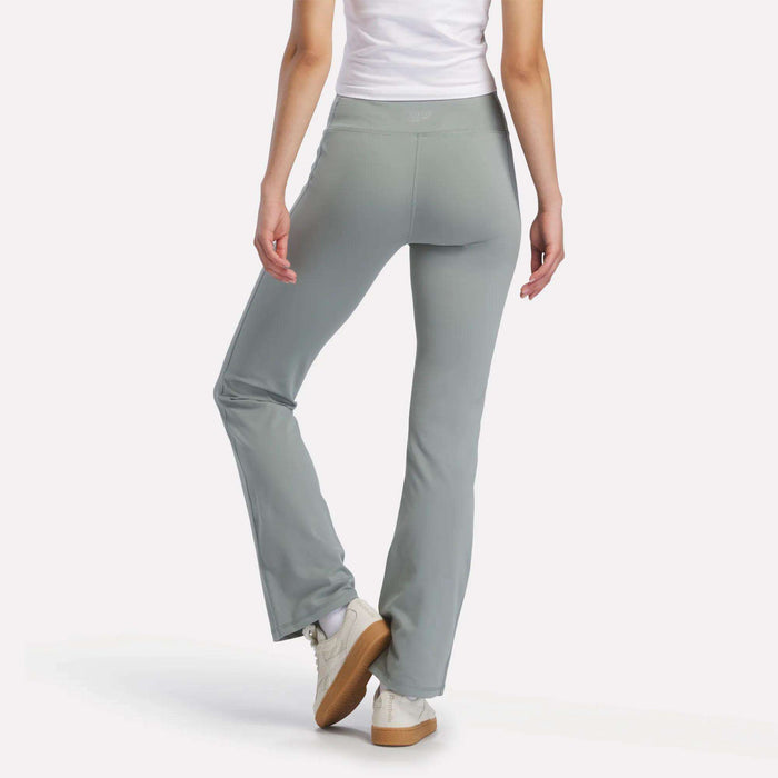 A woman stands with her back to the camera against a plain light background, wearing Reebok Women's Highstreet High-Rise Pants in light gray, paired with a white fitted t-shirt and white sneakers.