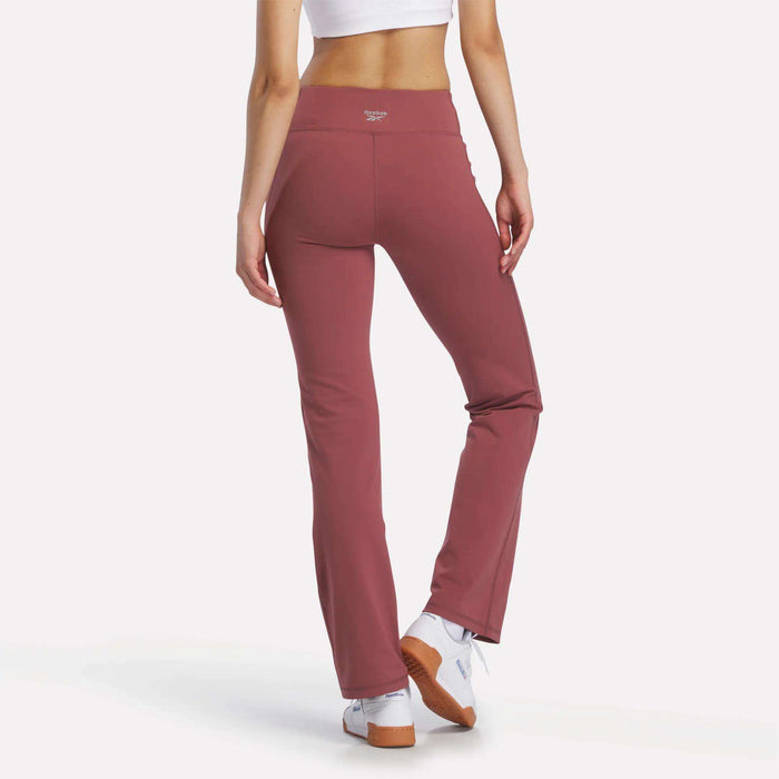 A person models Reebok Women's Highstreet High-Rise Pants in mauve, showing the back view with a white crop top and sneakers. The stretchy pants stand out against a plain white background.