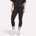 A figure from the shoulders down wears a loose black T-shirt, white sneakers, and Reebok Women's ID Basketball 3/4 Compression Leggings against a plain white background, highlighting the comfort and fit of these leggings.