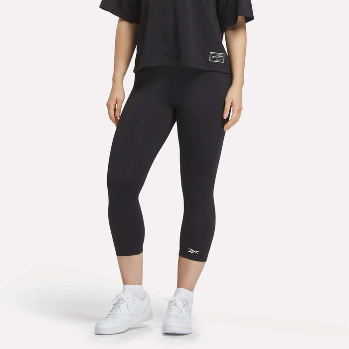 A figure from the shoulders down wears a loose black T-shirt, white sneakers, and Reebok Women's ID Basketball 3/4 Compression Leggings against a plain white background, highlighting the comfort and fit of these leggings.