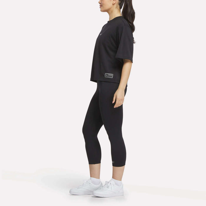 A woman wearing a black oversized t-shirt, Reebok Women's ID Basketball 3/4 Compression Leggings, and white sneakers stands sideways against a plain white background with her hair in a ponytail.