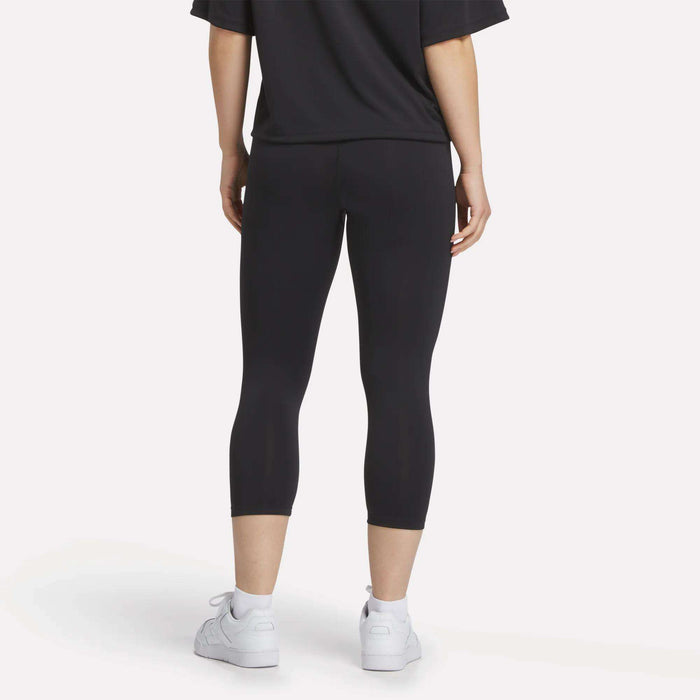 A person stands with their back to the camera, wearing a black t-shirt, white sneakers, and Reebok Women's ID Basketball 3/4 Compression Leggings against a plain light background.