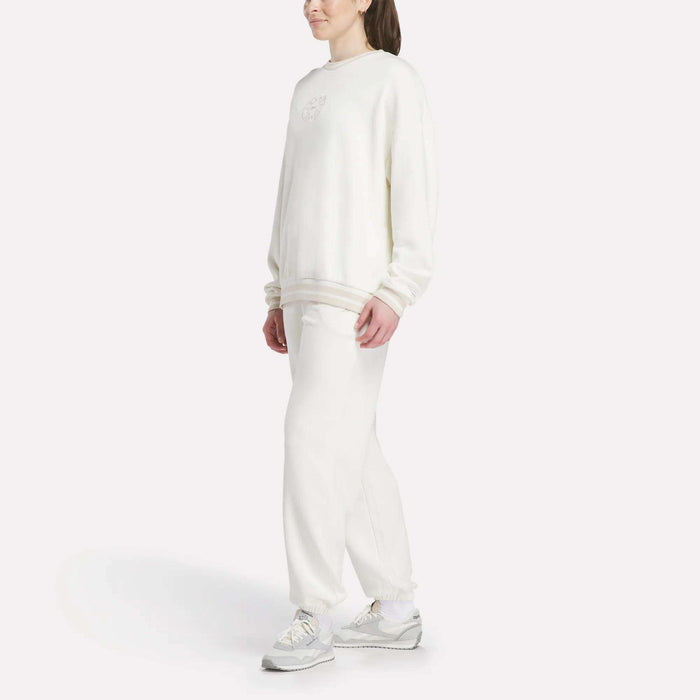 A person stands against a white background, wearing a loose-fitting Reebok Women's Lawn Club Crew Sweatshirt with matching white sweatpants and sneakers. Their hair is tied back, and their hands rest at their sides.