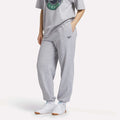 A person wearing a grey graphic t-shirt, Women's Lawn Club Jogger Pants by Reebok in light grey with a logo on the left thigh, and white sneakers, stands with hands in pockets against a plain background.