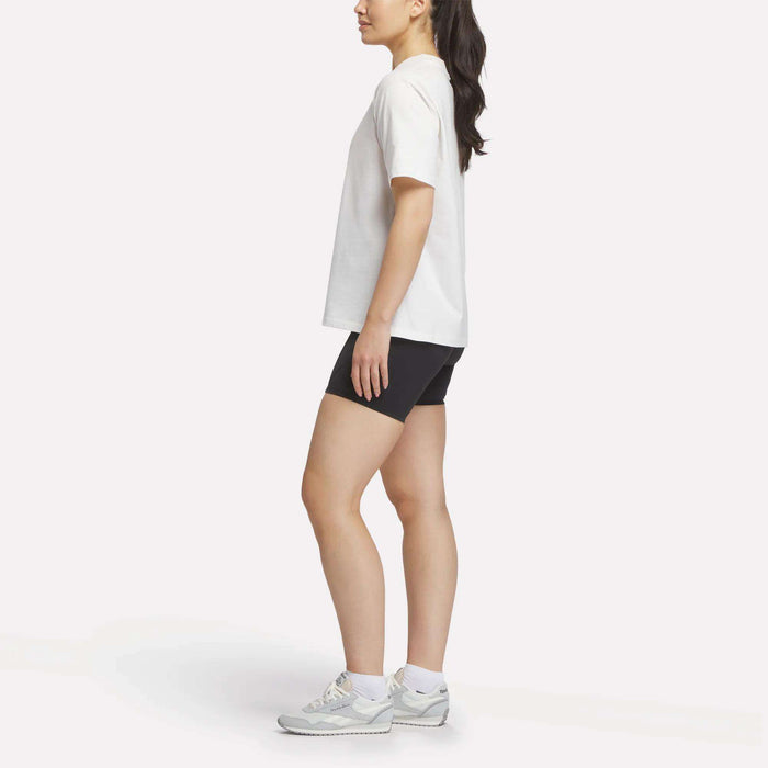 A woman stands in profile wearing the Reebok Women's Lawn Club T-Shirt, black shorts, white socks, and gray sneakers against a plain white background.