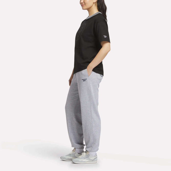 Woman wearing a Reebok Women's Lawn Club T-Shirt with a striped collar, grey Reebok sweatpants, and light grey sneakers, posed sideways against a white background. This tee offers casual comfort and sporty style.