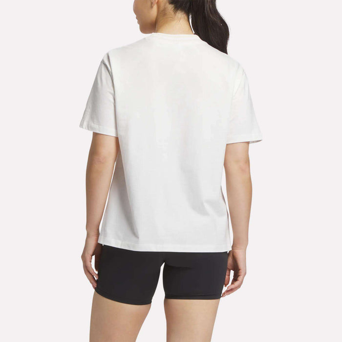 A woman with long dark hair in a ponytail is seen from behind, wearing the Reebok Women's Lawn Club T-Shirt in white and black shorts, set against a light background.
