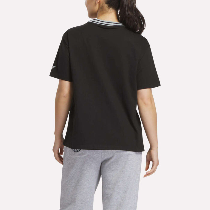 A woman with long dark hair in a ponytail is seen from behind, wearing a black Reebok Women's Lawn Club T-Shirt with a striped collar and light gray sweatpants, standing against a plain white background.