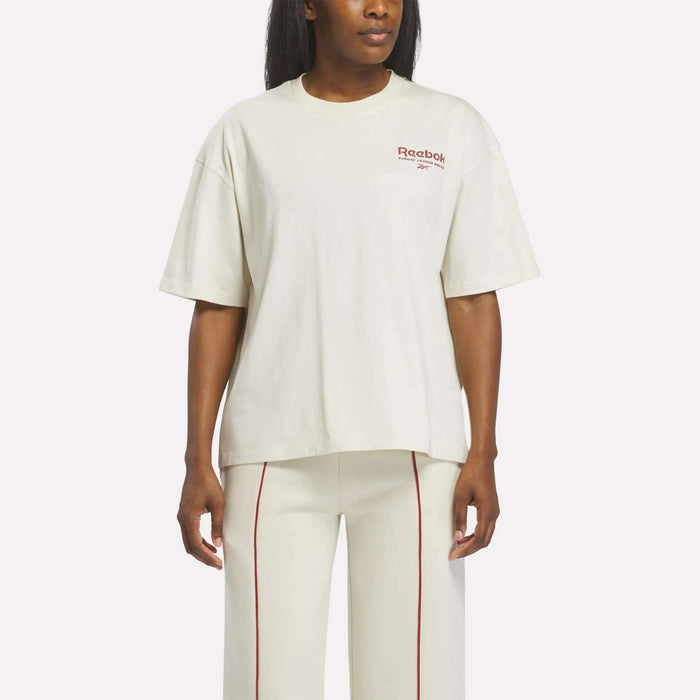 A woman stands against a plain background wearing a Reebok Women's League Club Graphic T-Shirt in linen and matching pants with red vertical stitching.