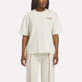 A woman stands against a plain background wearing a Reebok Women's League Club Graphic T-Shirt in linen and matching pants with red vertical stitching.