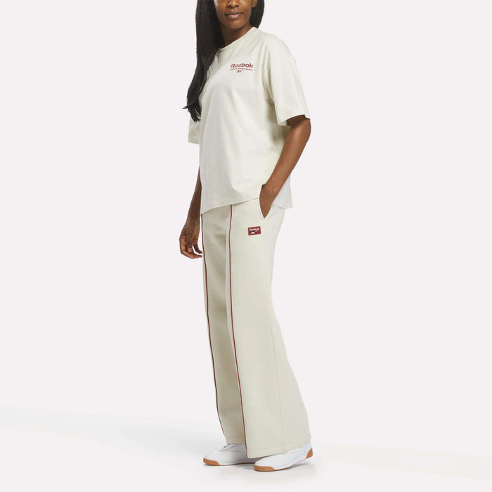 A person stands against a plain white background wearing a cream Women's League Club Graphic T-Shirt by Reebok with matching wide-leg pants featuring red trim and white sneakers.