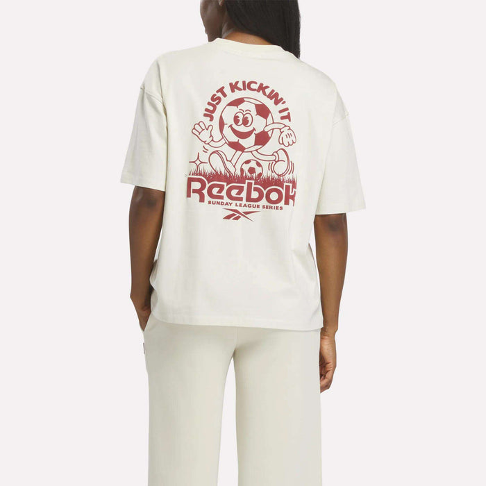 A person is seen from behind wearing the Reebok Women's League Club Graphic T-Shirt with a vintage soccer design and "JUST KICKIN IT" text, styled with matching white pants.