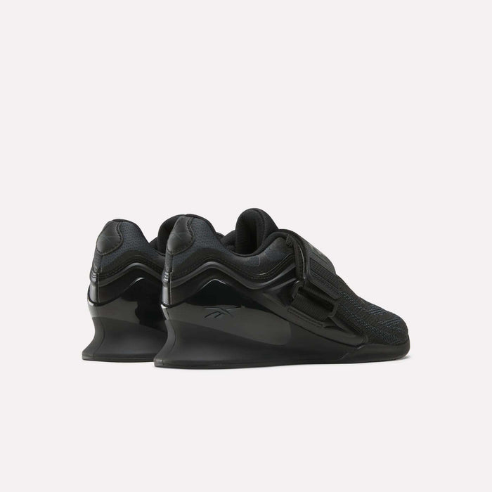 The Reebok Women's Legacy Lifter IV Training Shoes feature elevated heels, mesh uppers, wide straps, and glossy accents, shown from the back and side on a plain white background for support and stability.