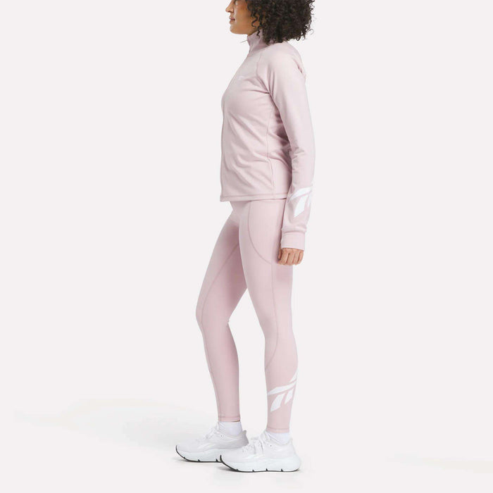A woman stands in profile against a plain white background, wearing Reebok Women's High-Rise Tights 26" in light pink with long sleeves and white sneakers.