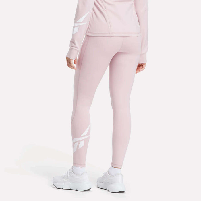 A person wears light pink Reebok Women's High-Rise Tights 26" with white graphic designs on the lower legs, a matching jacket, and white sneakers, standing with their back to the camera.
