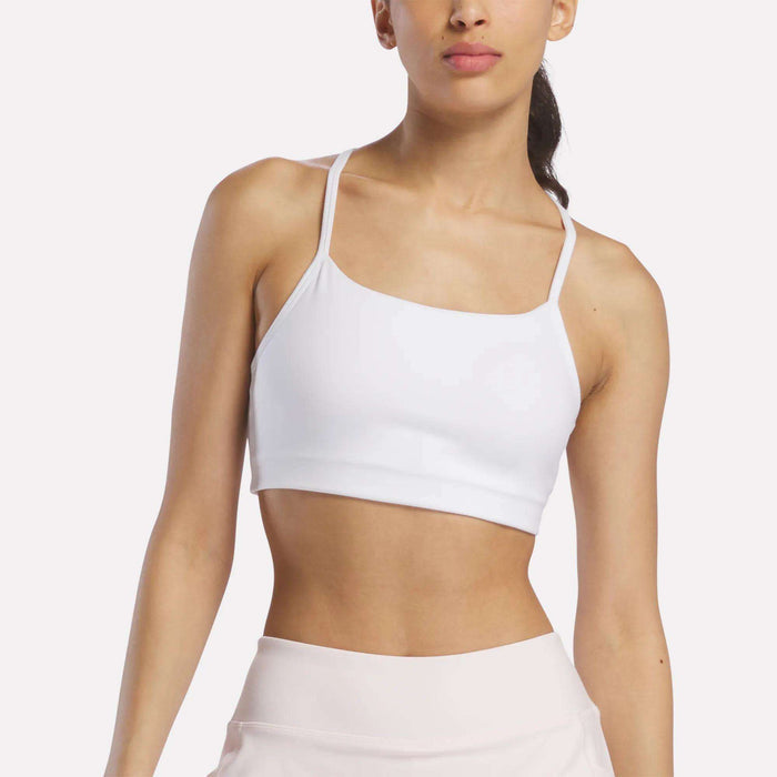 A woman models the Reebok Women's Low-Impact Sports Bra with adjustable straps, paired with light athletic pants. Posed athletically against a plain background, she showcases the ideal look for light-impact training.