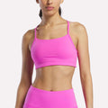 A woman models the Reebok Women's Low-Impact Sports Bra with adjustable straps in bright pink, paired with matching high-waisted leggings, against a plain white background. The image crops from her shoulders to just above her knees.