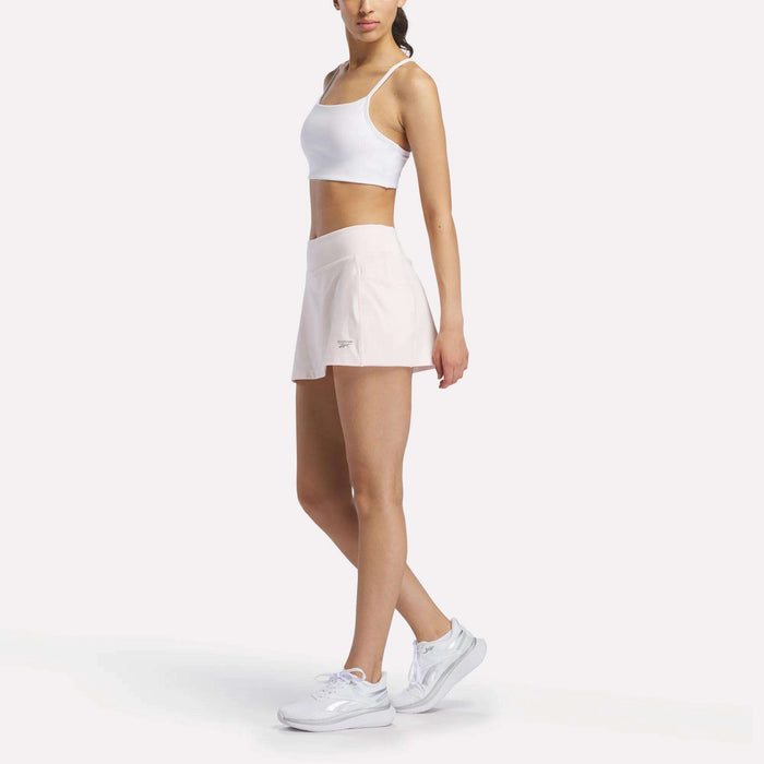 A woman stands against a white background wearing the Reebok Women's Low-Impact Sports Bra, paired with pale pink athletic shorts and white sneakers. She faces slightly to the side with one hand on her hip, ready for light-impact training.