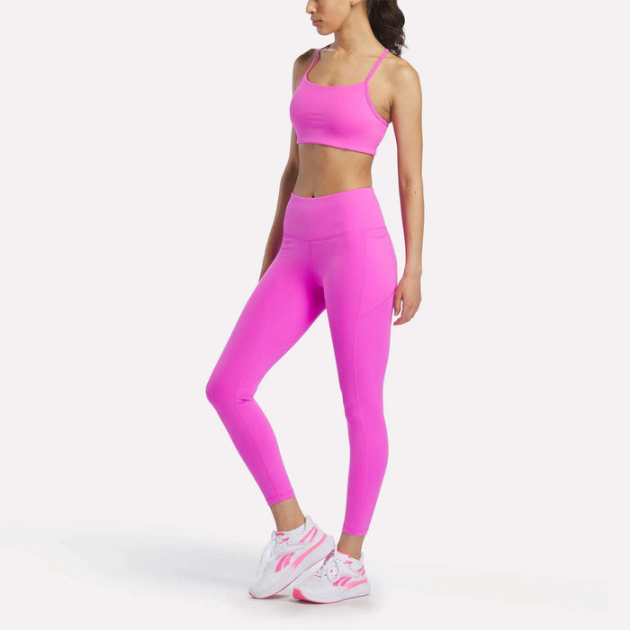 A woman poses in a three-quarter stance wearing the Reebok Women's Low-Impact Sports Bra and matching high-waisted leggings with white and pink sneakers, perfect for light-impact training, set against a plain white background.