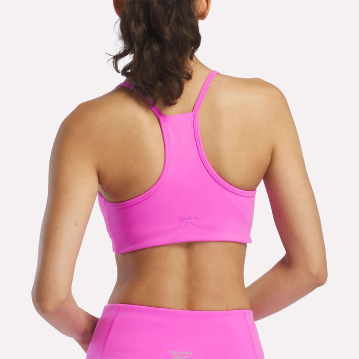 A woman with dark hair in a ponytail is seen from the back wearing Reebok’s Women's Low-Impact Sports Bra and matching leggings, both in bright pink with small Reebok logos on the back.