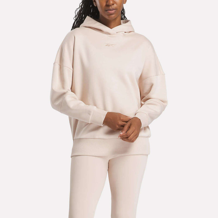 A woman in a light beige Reebok Women's Lux Hoodie and matching leggings stands with her hands clasped. The hoodie features a subtle chest logo, set against a plain white background.
