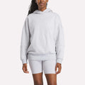 A woman wearing a cozy Reebok Women's Lux Hoodie in light gray and matching fitted shorts stands against a plain white background.