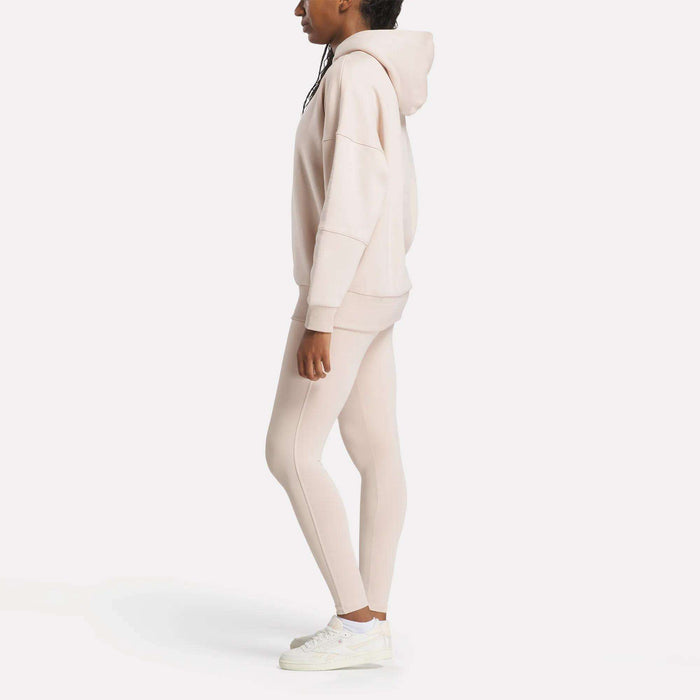 A person stands sideways against a white background, wearing a cozy Reebok Women's Lux Hoodie in beige, matching leggings, and white sneakers.