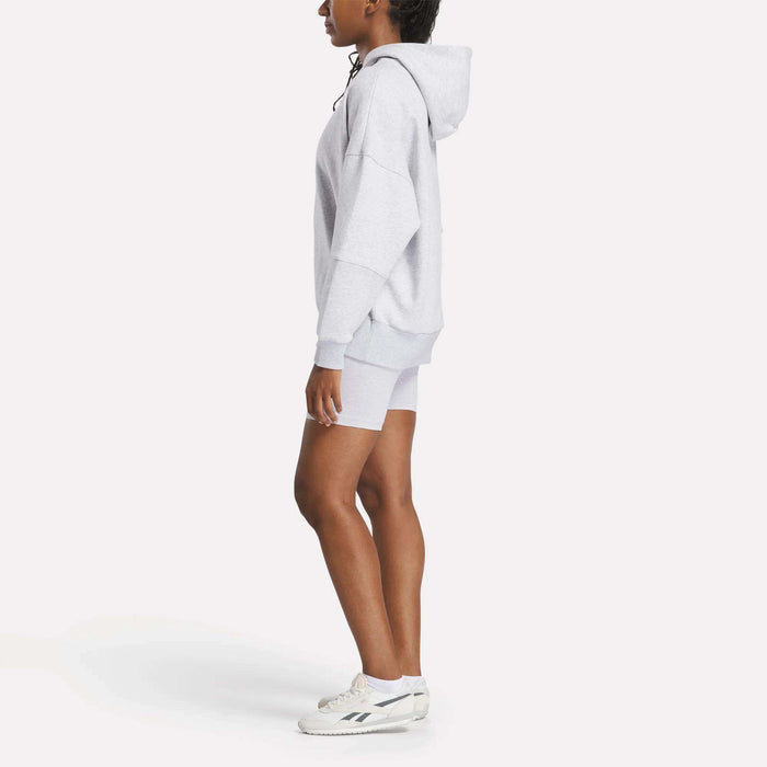 A person stands in profile against a plain white background, wearing a cozy Reebok Women's Lux Hoodie, matching gray shorts, and white sneakers.