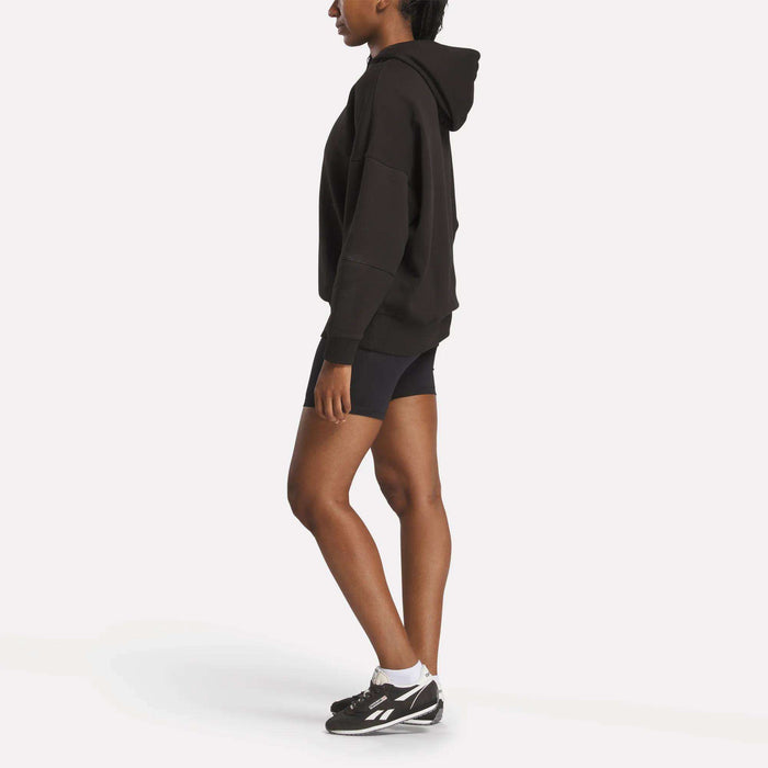 A woman stands sideways in a comfy Reebok Women's Lux Hoodie, paired with black athletic shorts, white socks, and black sneakers against a plain white background.