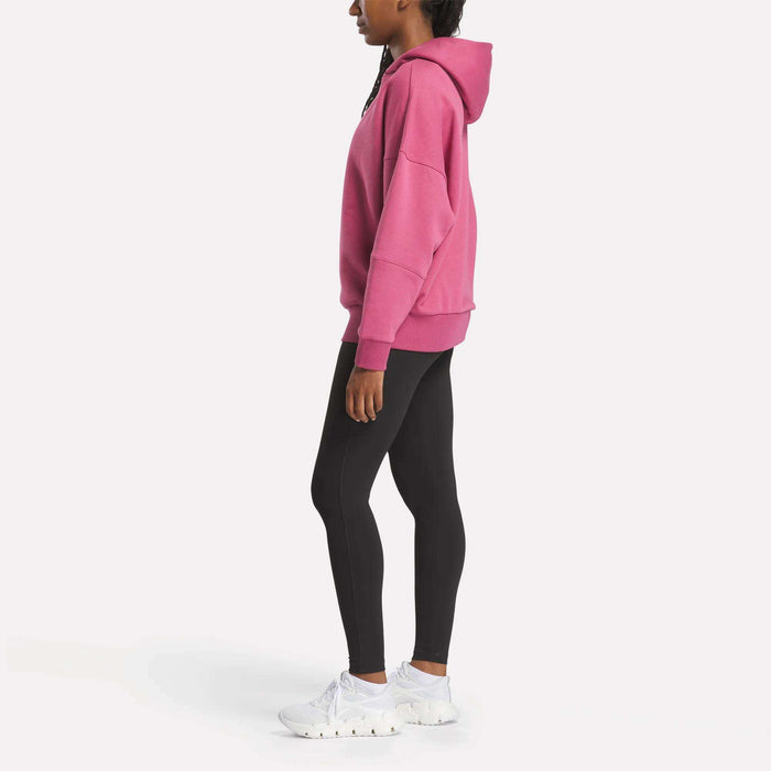 A woman stands in profile against a white background wearing the Reebok Women's Lux Hoodie in pink, paired with black leggings and white sneakers.