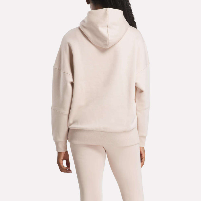 A woman with a ponytail is seen from the back wearing a light beige Women's Lux Hoodie by Reebok and matching fitted pants, standing against a plain white background with her left arm relaxed at her side.