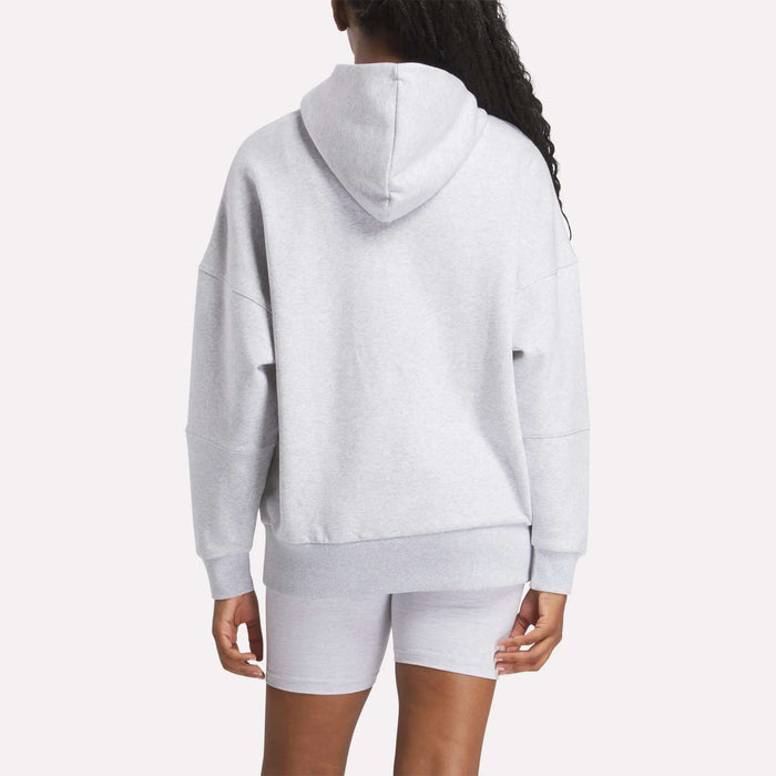 A woman wearing a Reebok Women's Lux Hoodie and matching shorts stands with her back to the camera against a plain light background, highlighting the cozy look of this light gray Reebok outfit.