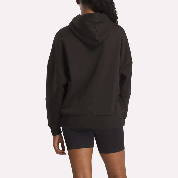 A woman with long curly hair, wearing the Reebok Women's Lux Hoodie and black fitted shorts, stands facing away from the camera against a plain light background.