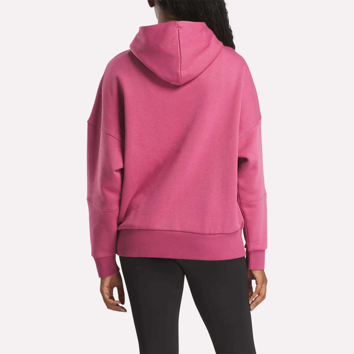 A person is seen from behind wearing a bright pink Reebok Women's Lux Hoodie and black pants, standing against a plain white background.