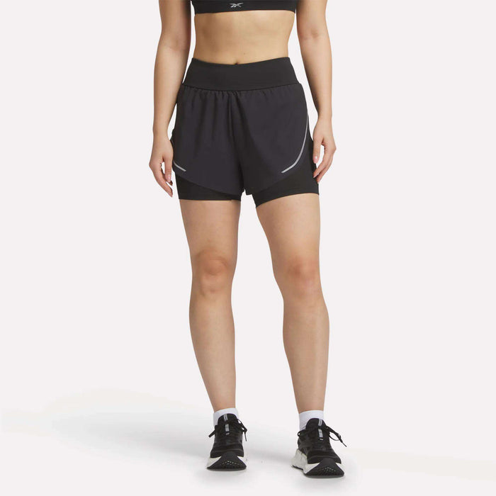 A person wears Reebok Women's Lux Metallic 2-in-1 Running Shorts 3", a black sports bra, white socks, and black running shoes, standing facing forward against a plain white background with their head out of frame.