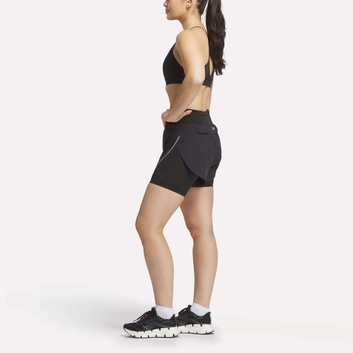 A woman in profile wears a black sports bra, Reebok Women's Lux Metallic 2-in-1 Running Shorts 3", white socks, and black athletic shoes against a plain white background.