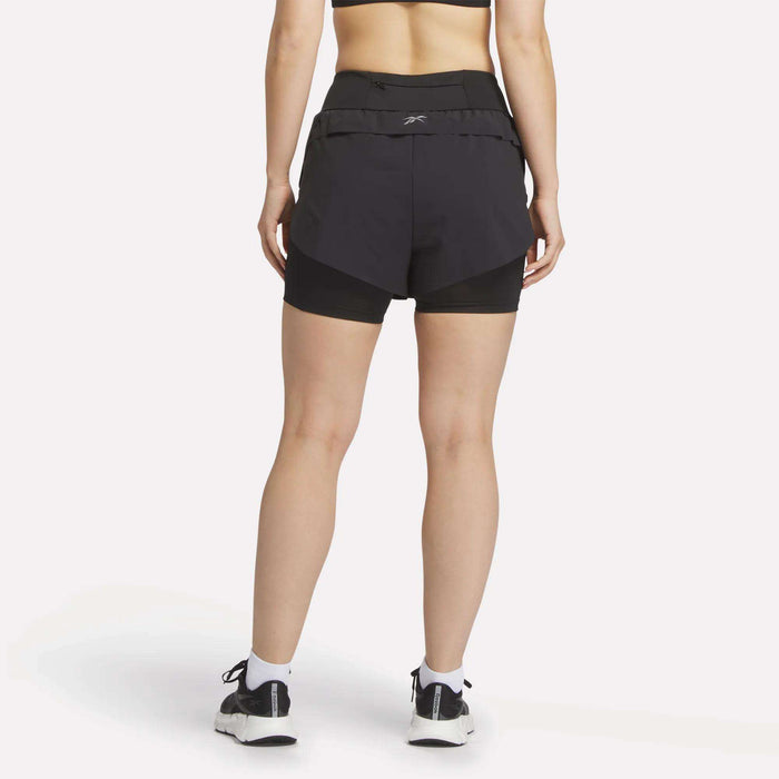 A woman faces away wearing Reebok Women's Lux Metallic 2-in-1 Running Shorts 3" with an elastic waistband, a black sports bra, white socks, and black and white sneakers against a plain white background.