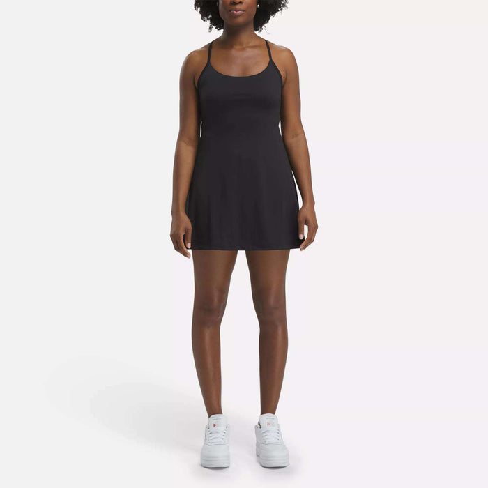 A woman faces forward against a plain white background, wearing the Reebok Women's Lux Strappy Dress in black with white sneakers.