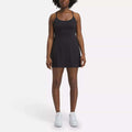 A woman faces forward against a plain white background, wearing the Reebok Women's Lux Strappy Dress in black with white sneakers.