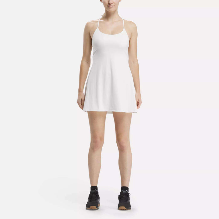 A woman faces forward wearing the Reebok Women's Lux Strappy Dress, a sleeveless sporty dress with moisture-wicking fabric and flared skirt, paired with black athletic shoes, all set against a plain white background.
