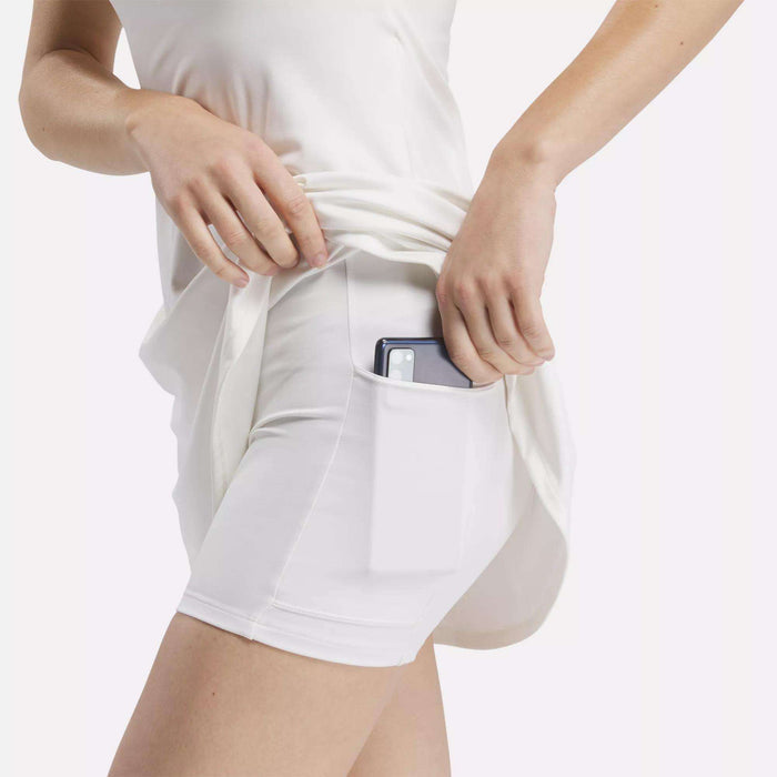 A woman wears the Reebok Women's Lux Strappy Dress in white, placing her smartphone into the side pocket of the moisture-wicking dress, with the pocket clearly visible.