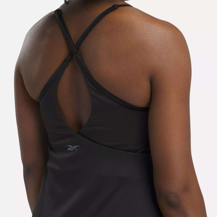 A woman wears the Reebok Women's Lux Strappy Dress featuring adjustable crisscross straps, a keyhole back, and visible branding. A plain background highlights its moisture-wicking fabric.