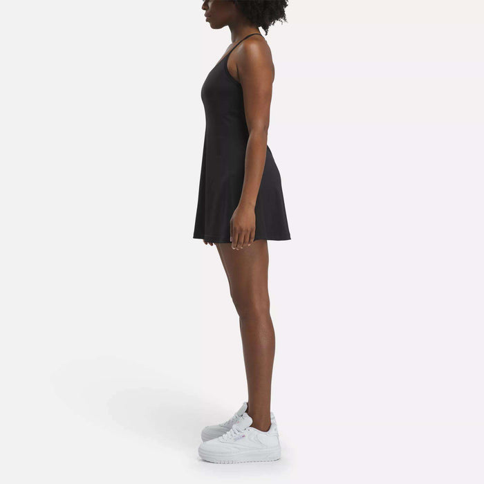 A woman stands in profile against a white background, wearing the Reebok Women's Lux Strappy Dress with adjustable straps and white sneakers.