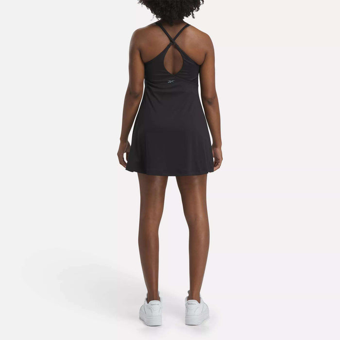 A person with curly hair faces away, wearing the Reebok Women's Lux Strappy Dress with adjustable straps and white sneakers, set against a plain, light background.