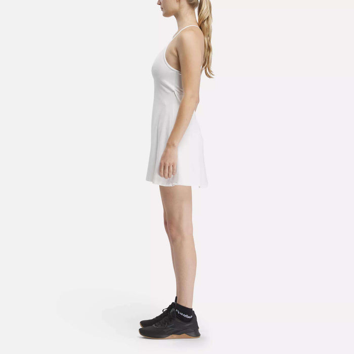 A woman with long blonde hair in a ponytail stands in profile, wearing the Reebok Women's Lux Strappy Dress with black athletic shoes and black socks against a plain white background.