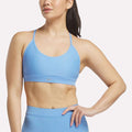 A woman models the Reebok Women's Lux Strappy Sports Bra in light blue, standing with one hand on her hip against a white background. The sweat-wicking fabric keeps her comfortable and cool.