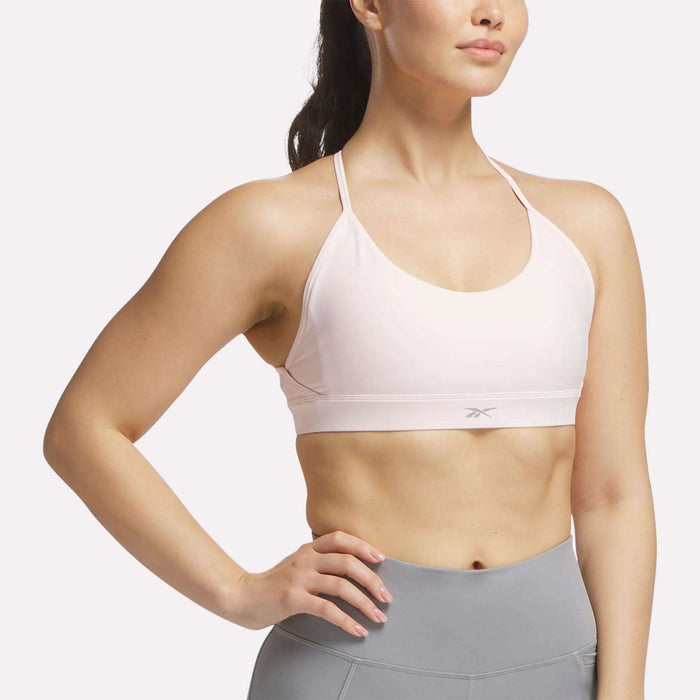 A woman in a Reebok Women's Lux Strappy Sports Bra (light pink) and high-waisted gray leggings stands with one hand on her hip, facing slightly sideways against a plain background.