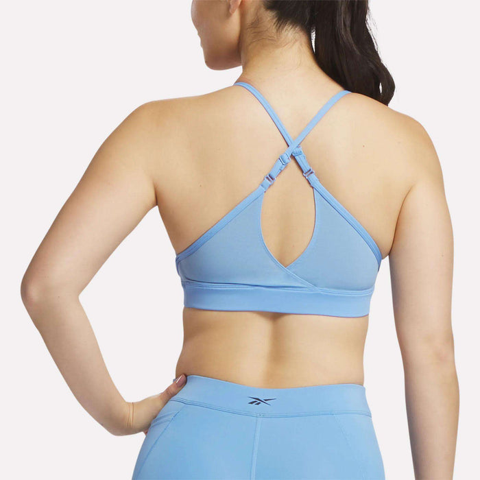 A woman is seen from the back wearing a Reebok Women's Lux Strappy Sports Bra in light blue with criss-cross straps, paired with matching leggings, set against a plain white background.